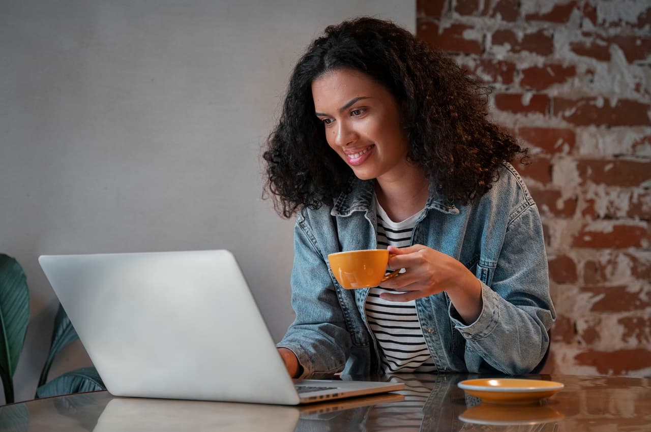 Mulher sorridente de cabelos cacheados médios, segura xícara de café amarela e digita no computador.