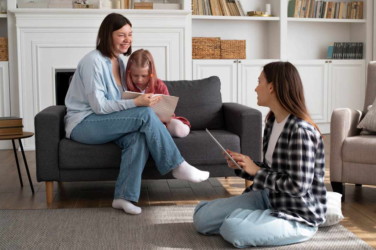 Família sorridente (duas mulheres e uma criança) lendo livros na sala de estar. Uma das mulheres abraça a criança, enquanto a outra está sentada no chão sorrindo para elas.