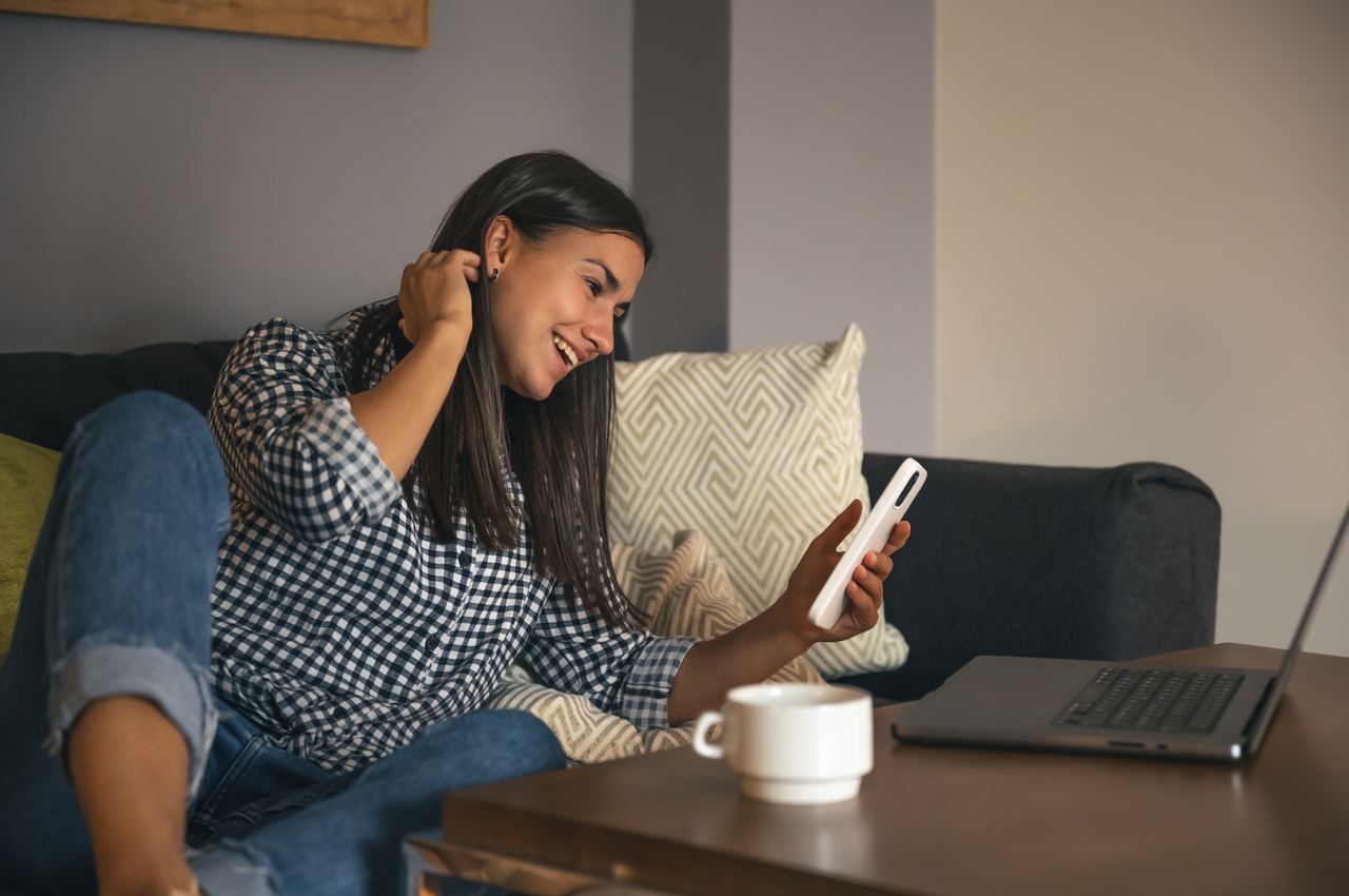 Mulher sorridente de cabelos castanhos longos, acessando boletos online pelo celular. Ela está sentada na cama e descalça, vestindo calça jeans e camisa básica