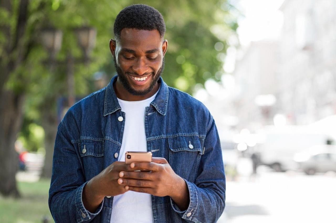 Homem sorridente digitando no celular no meio de uma praça arborizada. Ele veste conjunto jeans.