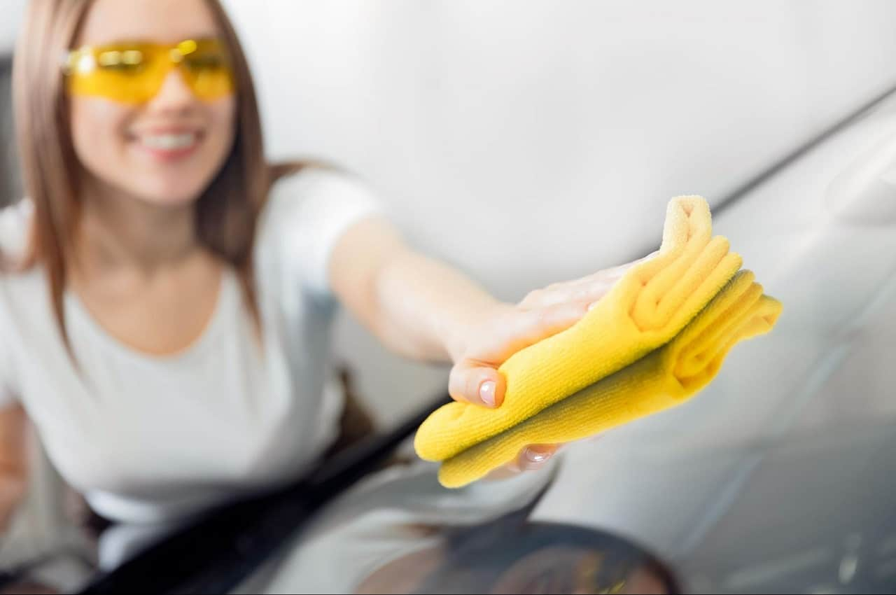 Mulher sorridente de cabelos castanhos médios usa óculos de proteção amarelo e flanela amarela para fazer a higienização de carros. 