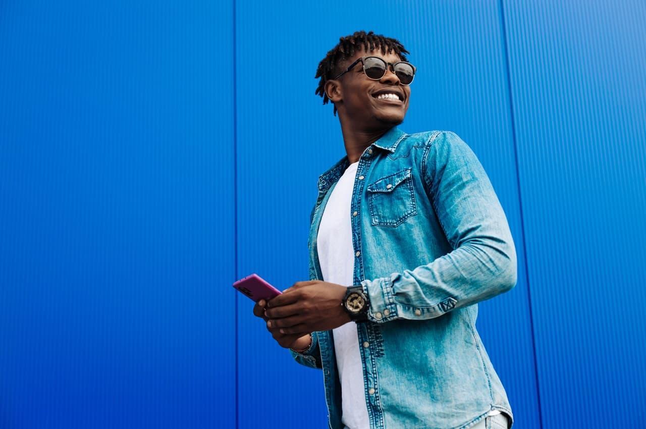 Homem sorridente de óculos escuros, segura celular com as duas mãos e anda ao lado de uma parede azul anil na rua. Ele veste camisa básica branca e jaqueta jeans.