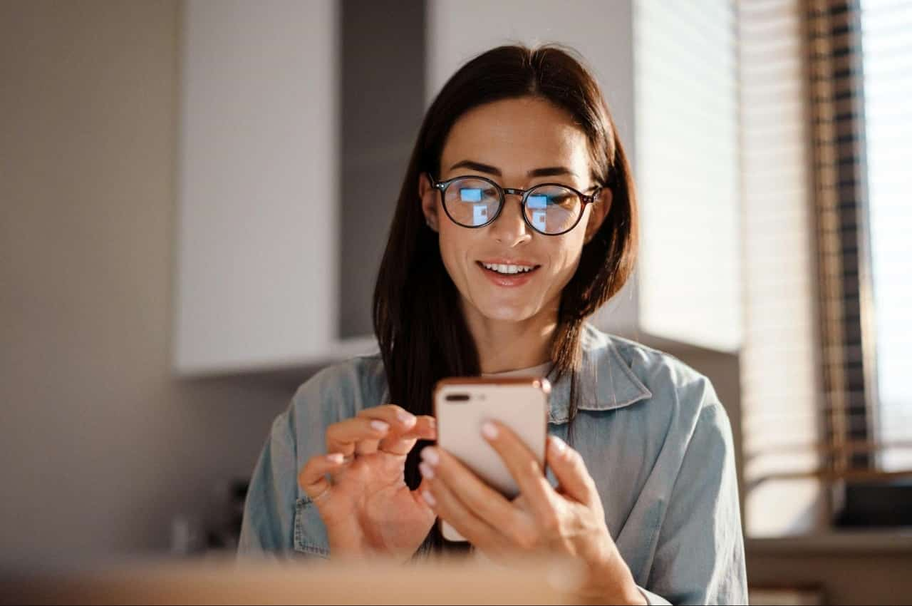 Mulher sorridente usando óculos de grau que reflete a tela do celular que ela utiliza na imagem.