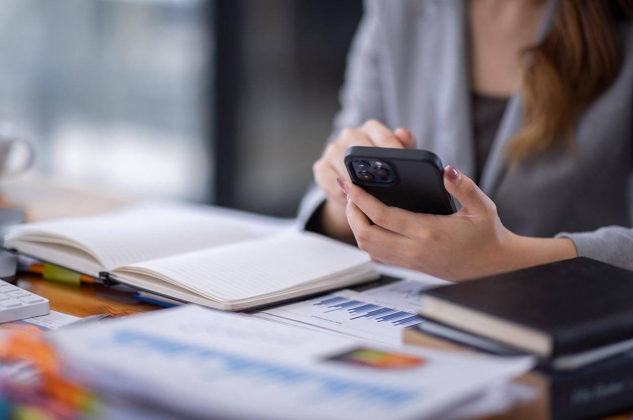 A imagem mostra uma mão usando um celular em uma mesa. Nela, contém alguns papéis, agendas e cadernetas.