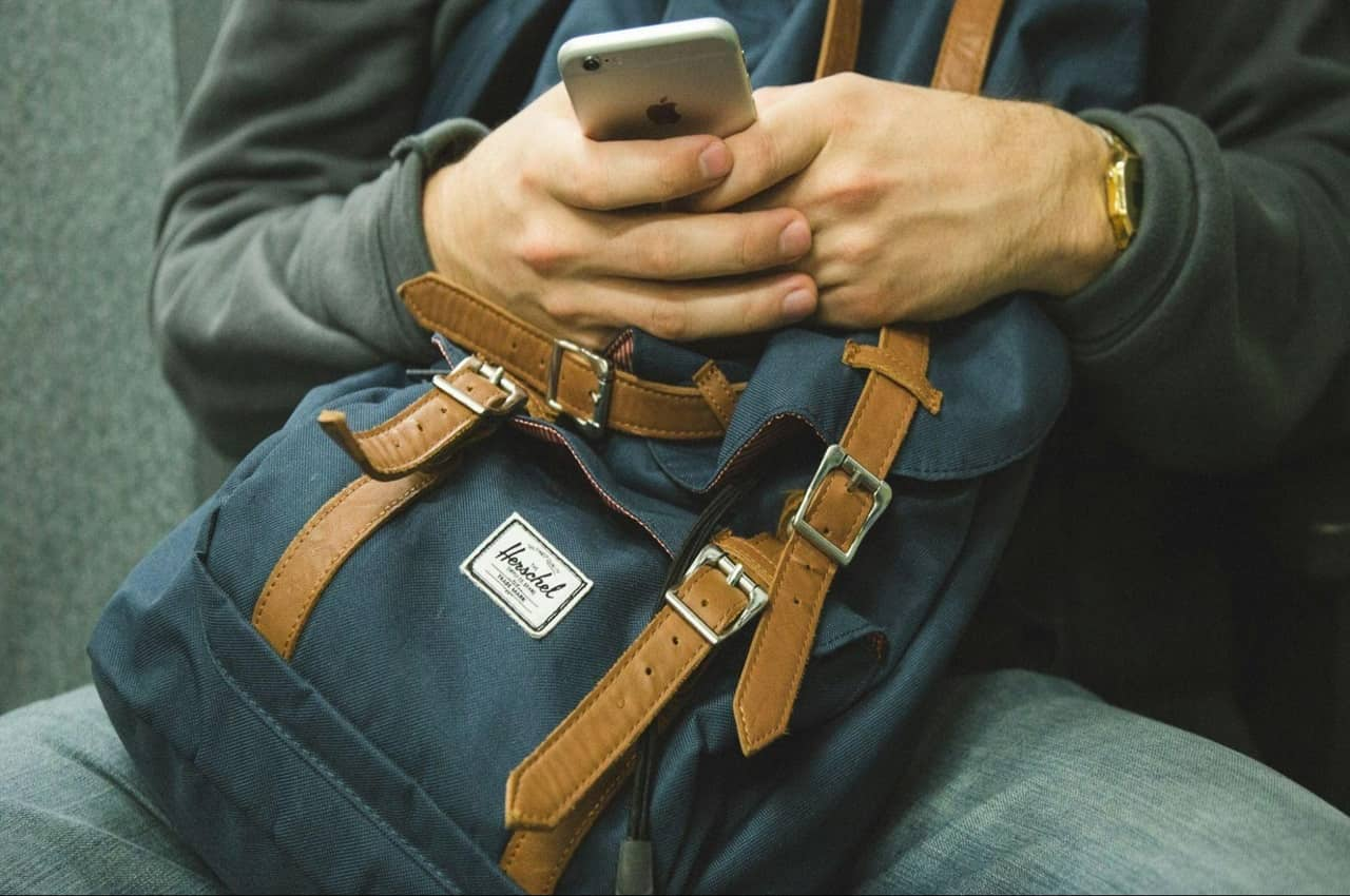 Mãos segurando um celular e uma mochila. O rosto da pessoa não aparece na imagem e ela está sentada em um banco.