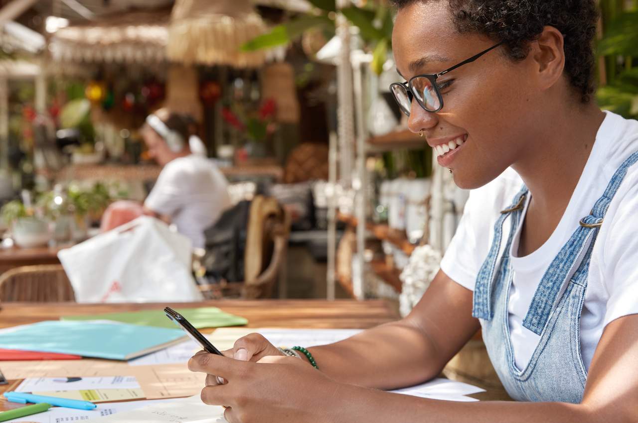 Mulher sorridente com óculos de grau e vestindo macacão jeans, pagando boletos online com celular. Tem vários papéis espalhados pela sua mesa.