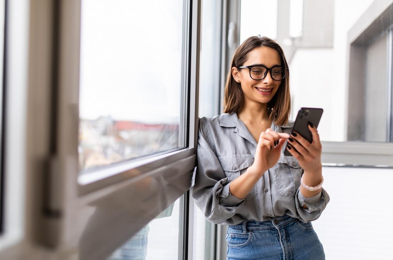 Mulher sorridente encostada no batente de uma janela fechada e digitando acessando consignado online pelo celular. Ela usa óculos de grau, camisa social cinza e calça jeans.