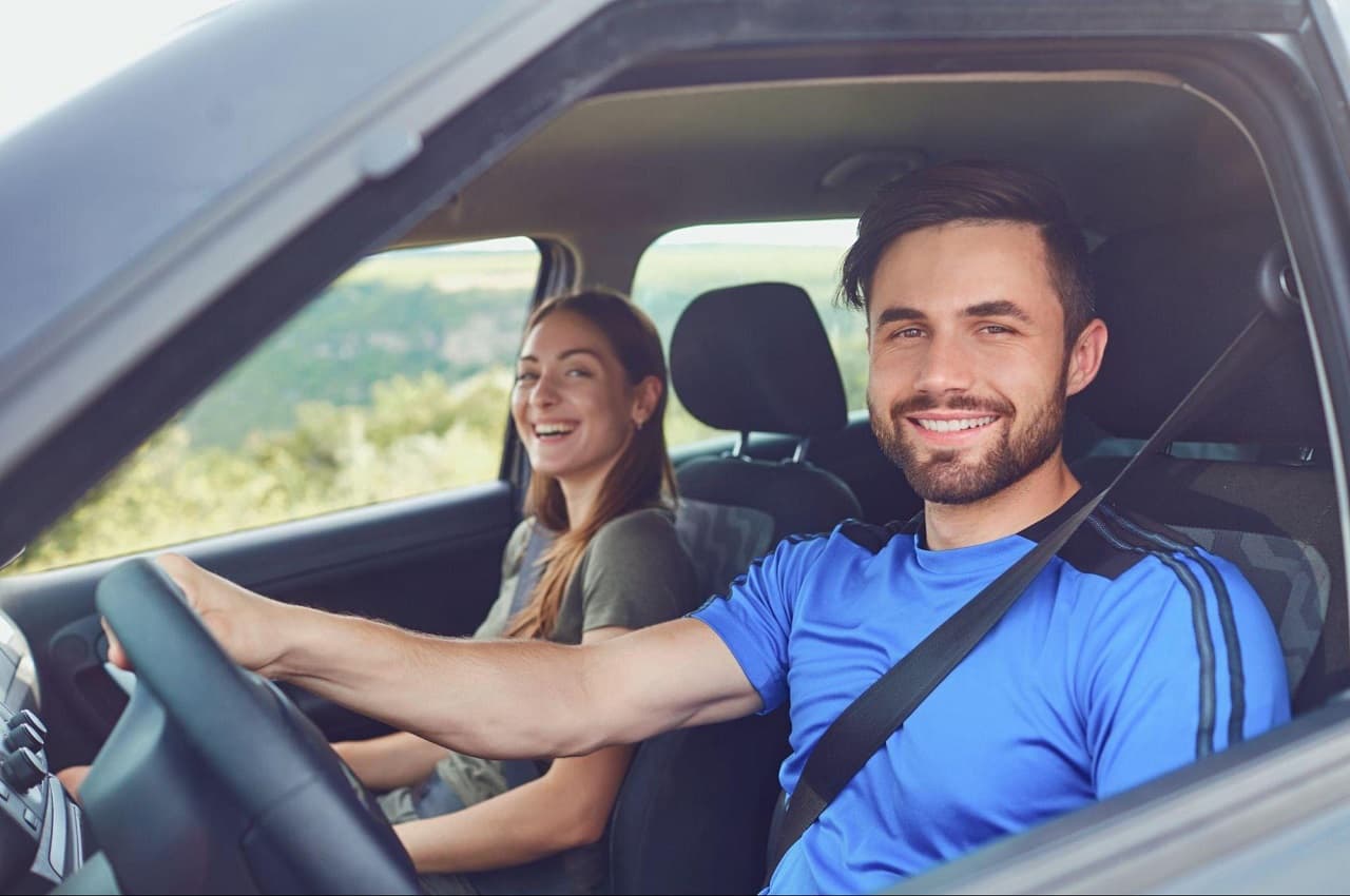 Homem sorridente, vestindo camisa azul anil e dirigindo um carro. No banco do passageiro, tem uma mulher de cabelos castanhos sorridente.