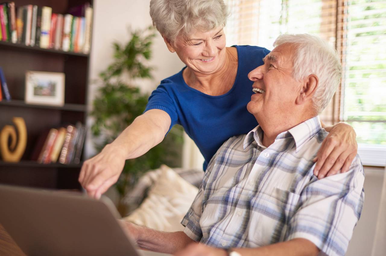 A imagem mostra um casal de idosos usando um laptop. Eles estão sorridentes.