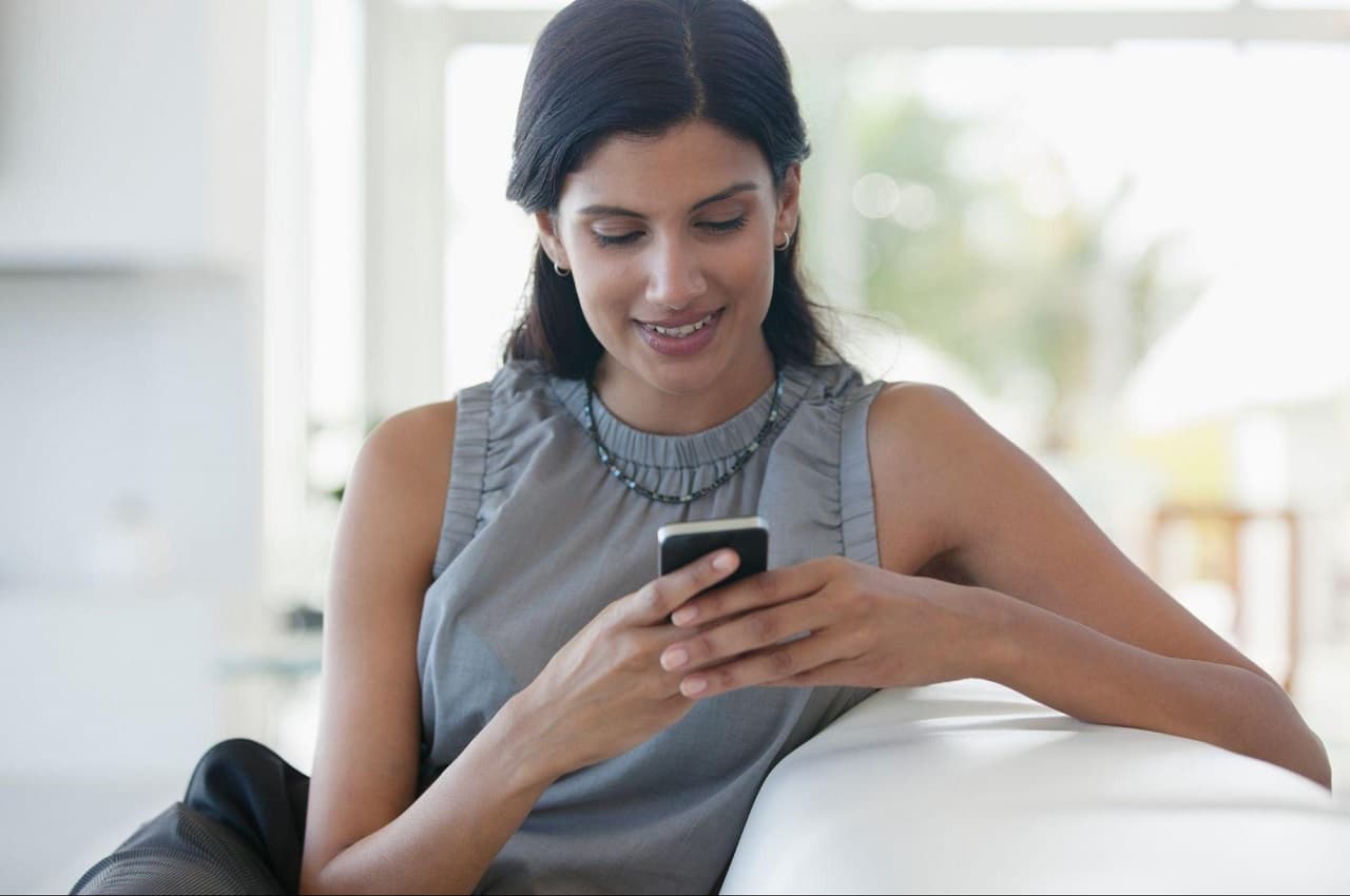 Mulher sorridente sentada em seu sofá e pesquisando como quitar dívidas no seu celular