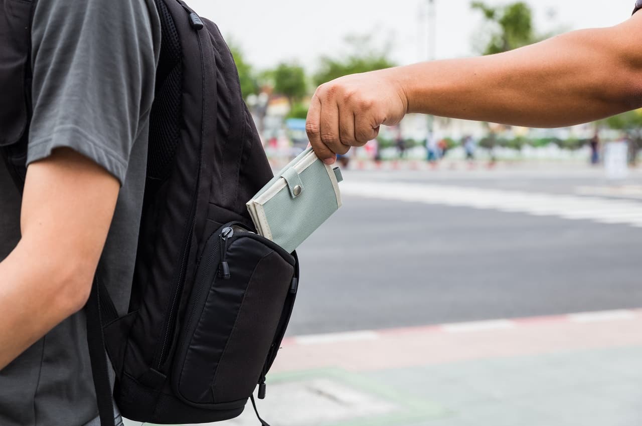 Pessoa (rosto não aparece na imagem) roubando carteira da mochila de um homem andando na rua