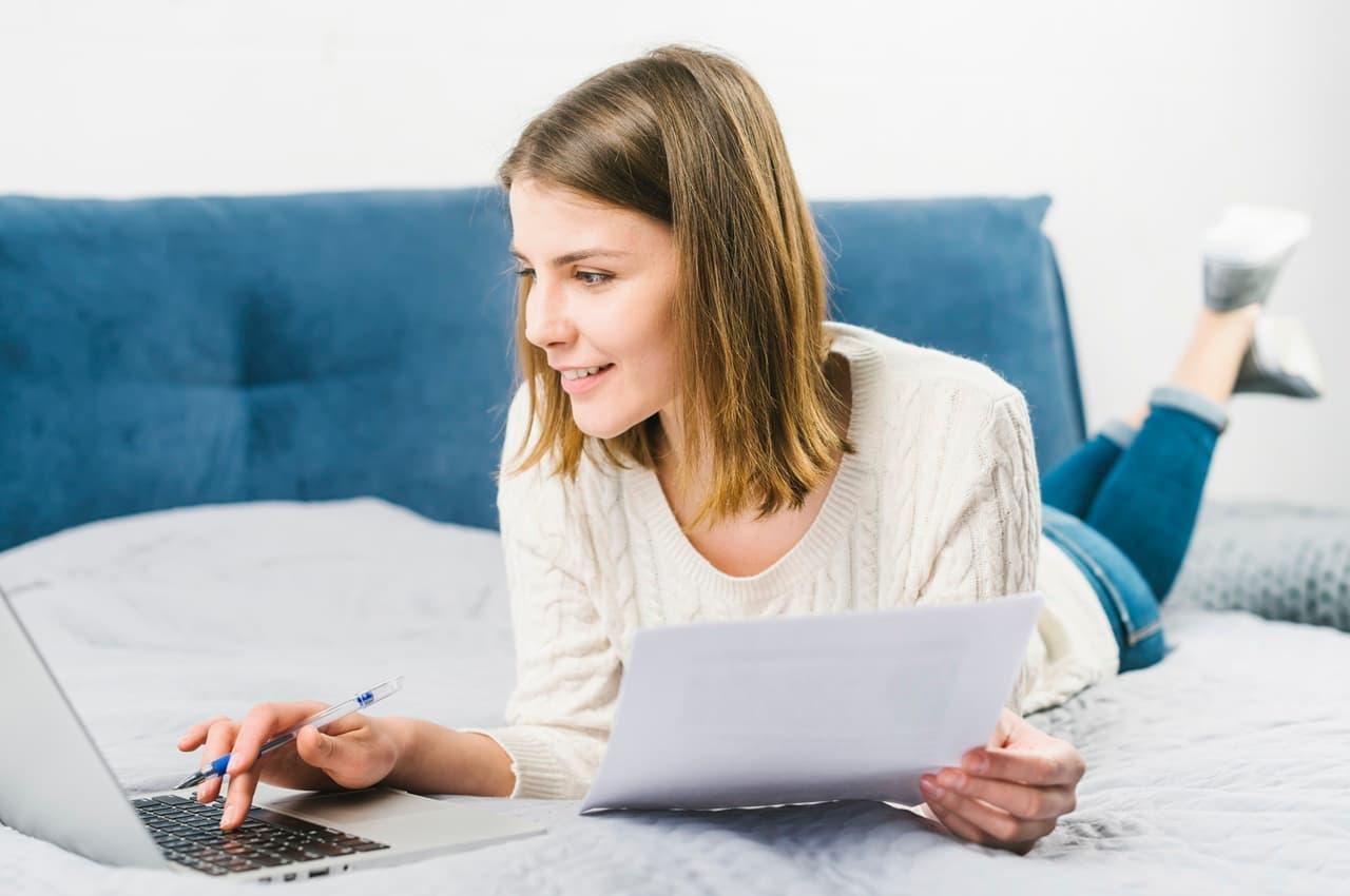 Mulher de cabelo liso, vestindo um suéter branco, deitada em uma cama enquanto usa um notebook. Ela segura papéis com uma mão e está concentrada, pesquisando sobre parcela de financiamento.