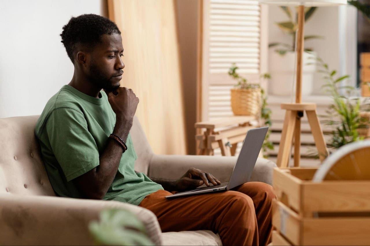 Homem com expressão concentrada sentado no sofá utilizando laptop para renegociar financiamento, Ele veste calça bege e camiseta verde.