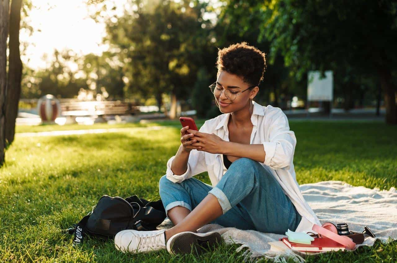 Mulher sorridente de óculos de grau e usando um celular. Ela está sentada em cima de uma toalha de piquenique em um parque.