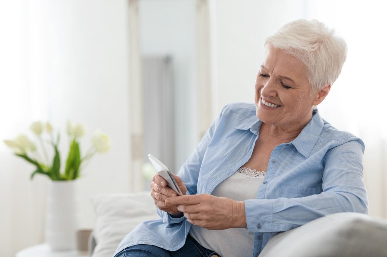 Mulher sorridente sênior de cabelos curtos grisalhos, sentada no sofá e digitando no celular.