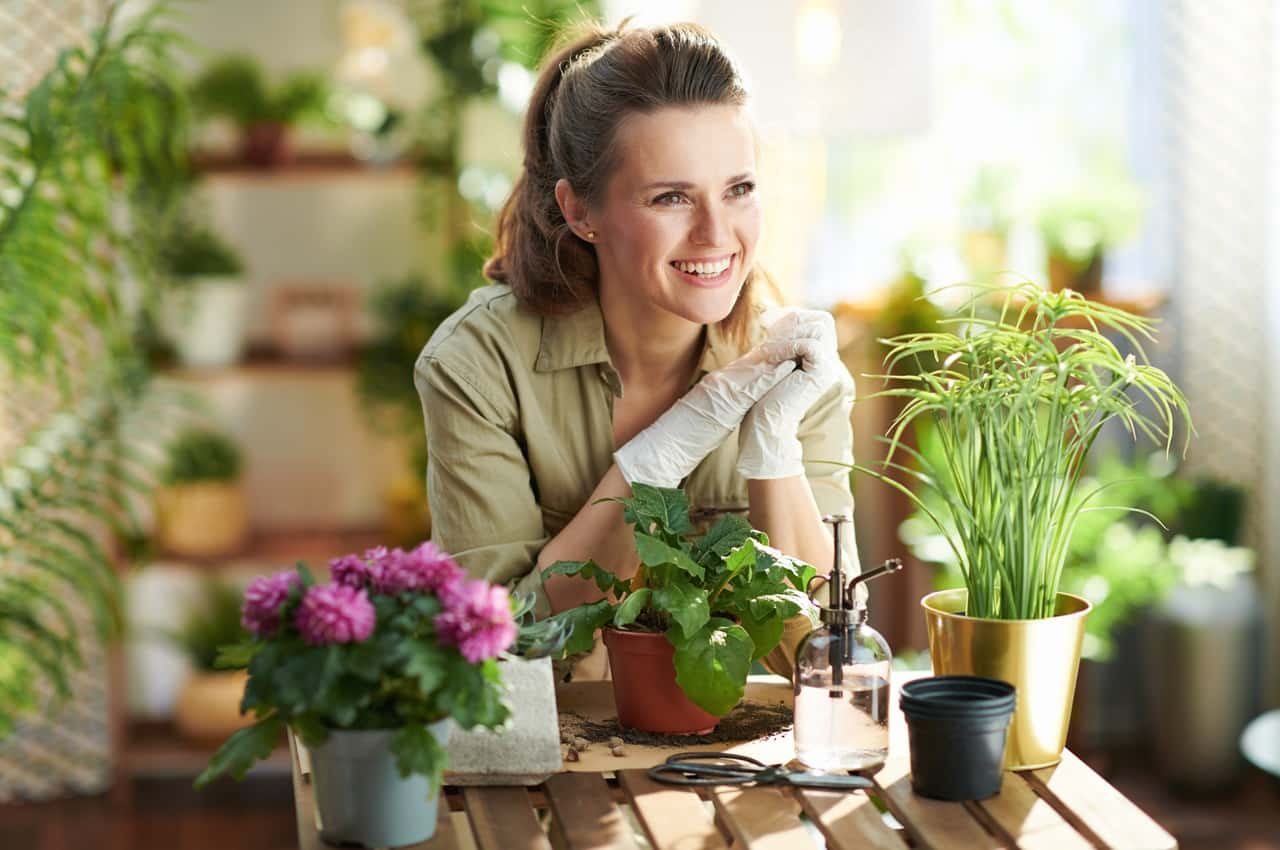 Mulher sorridente usando luvas de jardinagem, cuidando de uma casa sustentável com diferentes vasos de flores.