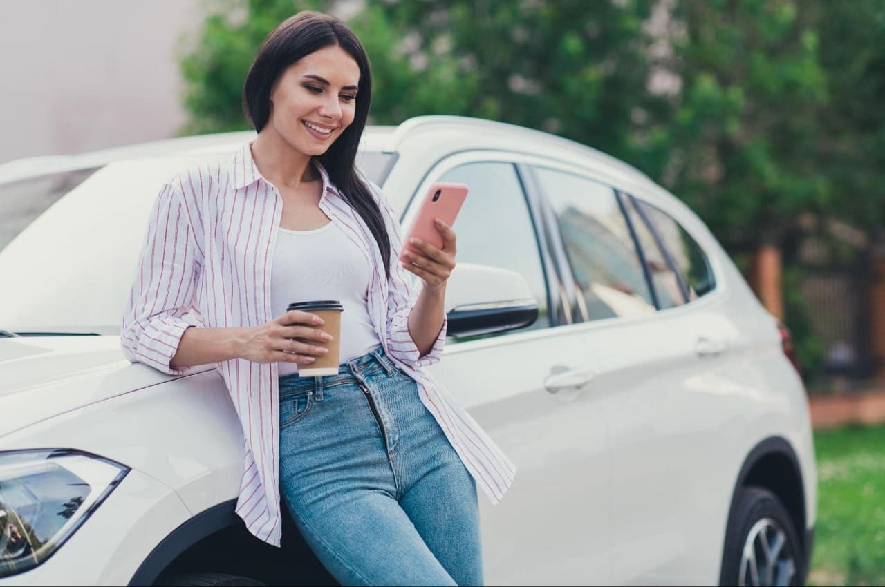 Mulher sorridente de cabelos castanhos, utilizando um celular e segurando um copo de café. Ela está apoiada na lateral de um carro branco.