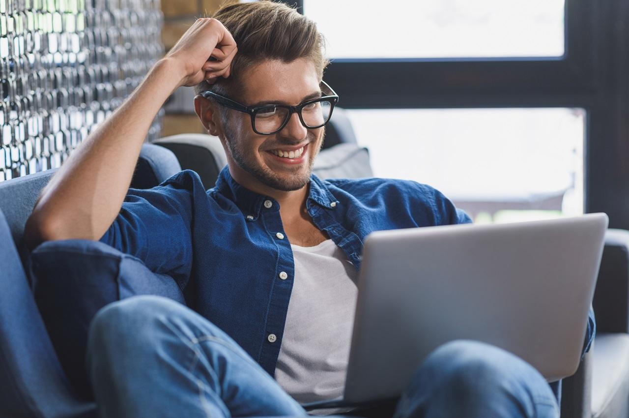 Homem sorridente de óculos de grau, usando um computador que esta em seu colo. O homem está sentado no sofá com uma mão apoiada sua cabeça.