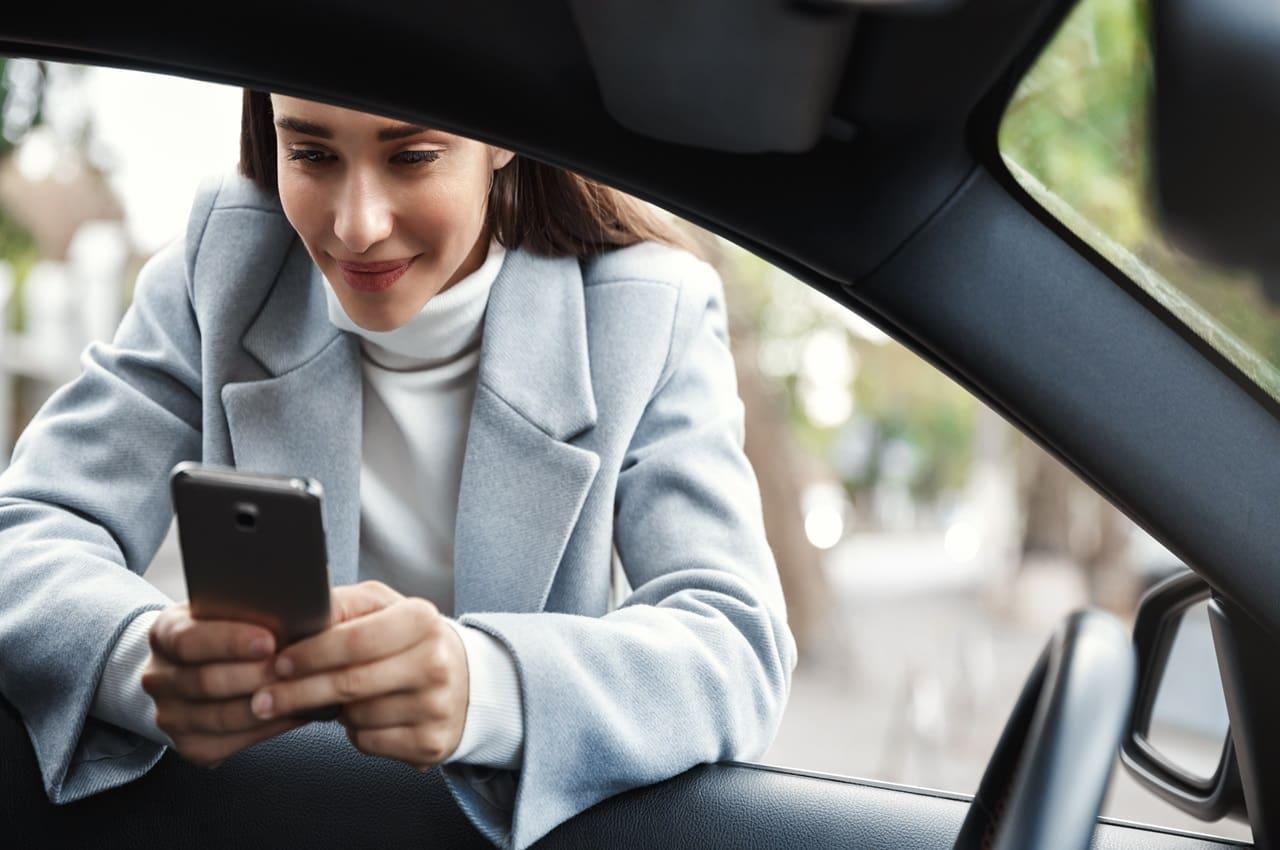 Mulher sorridente olhando para o celular enquanto se apoia na janela de um carro. Ela está pesquisando sobre como utilizar o Renave.