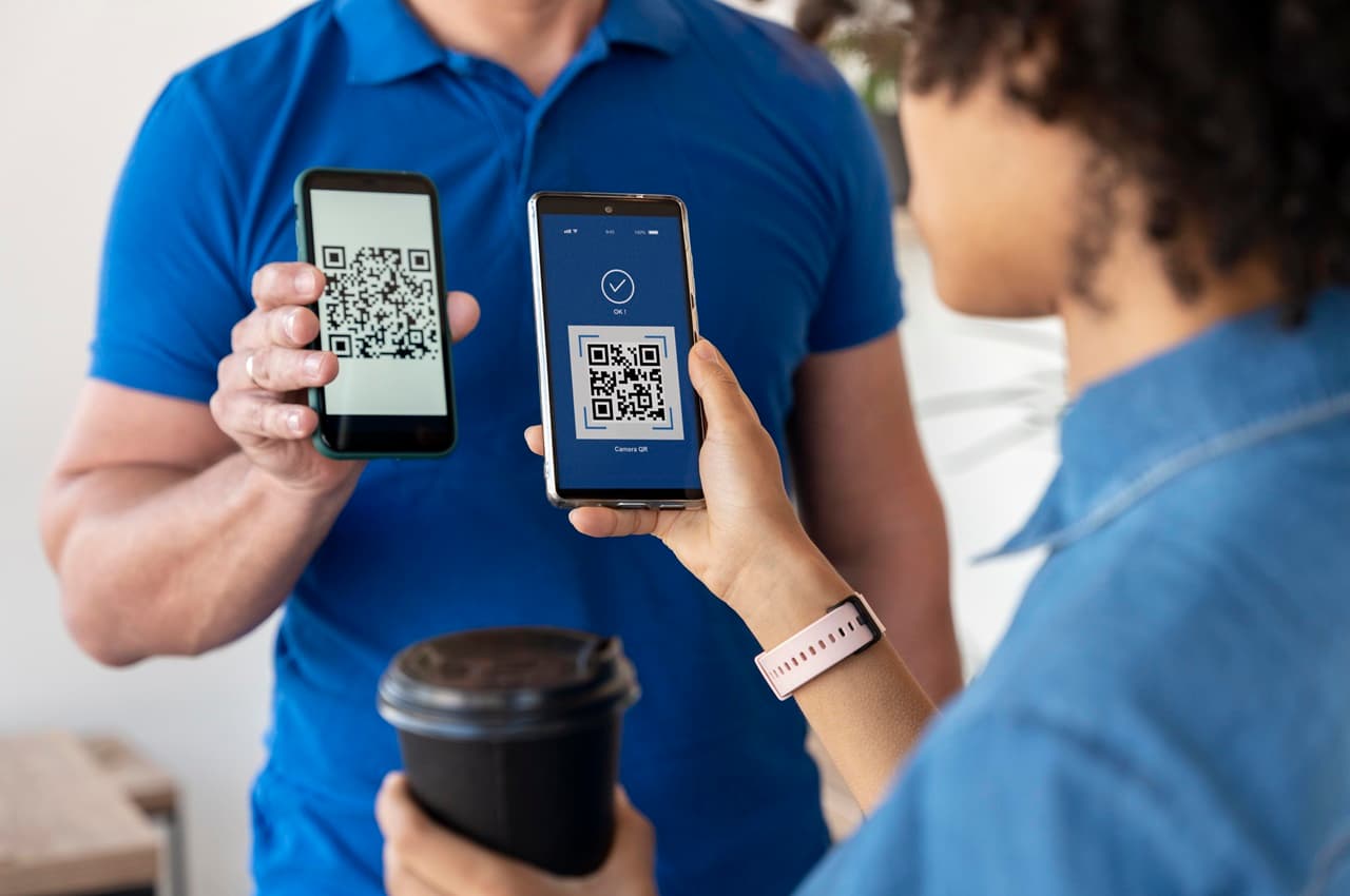 Atendente de camisa polo azul mostrando QR Code de pagamento para um homem que usa celular para pagar Pix.