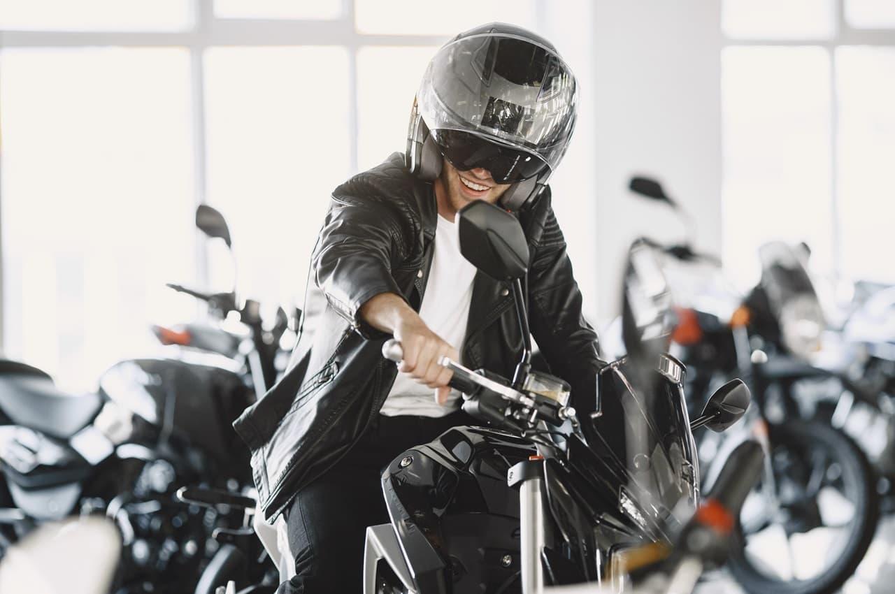 Homem comprando sua primeira moto em uma concessionária. Ele está com uma expressão feliz e com um capacete na cabeça.