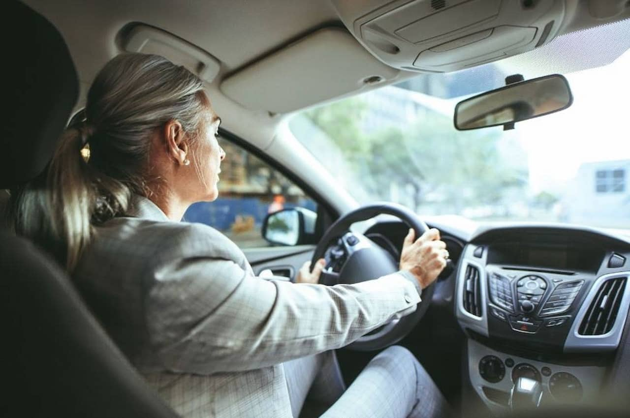 Mulher de cabelos grisalhos e expressão concentrada dirigindo seu veículo em um via asfaltada.