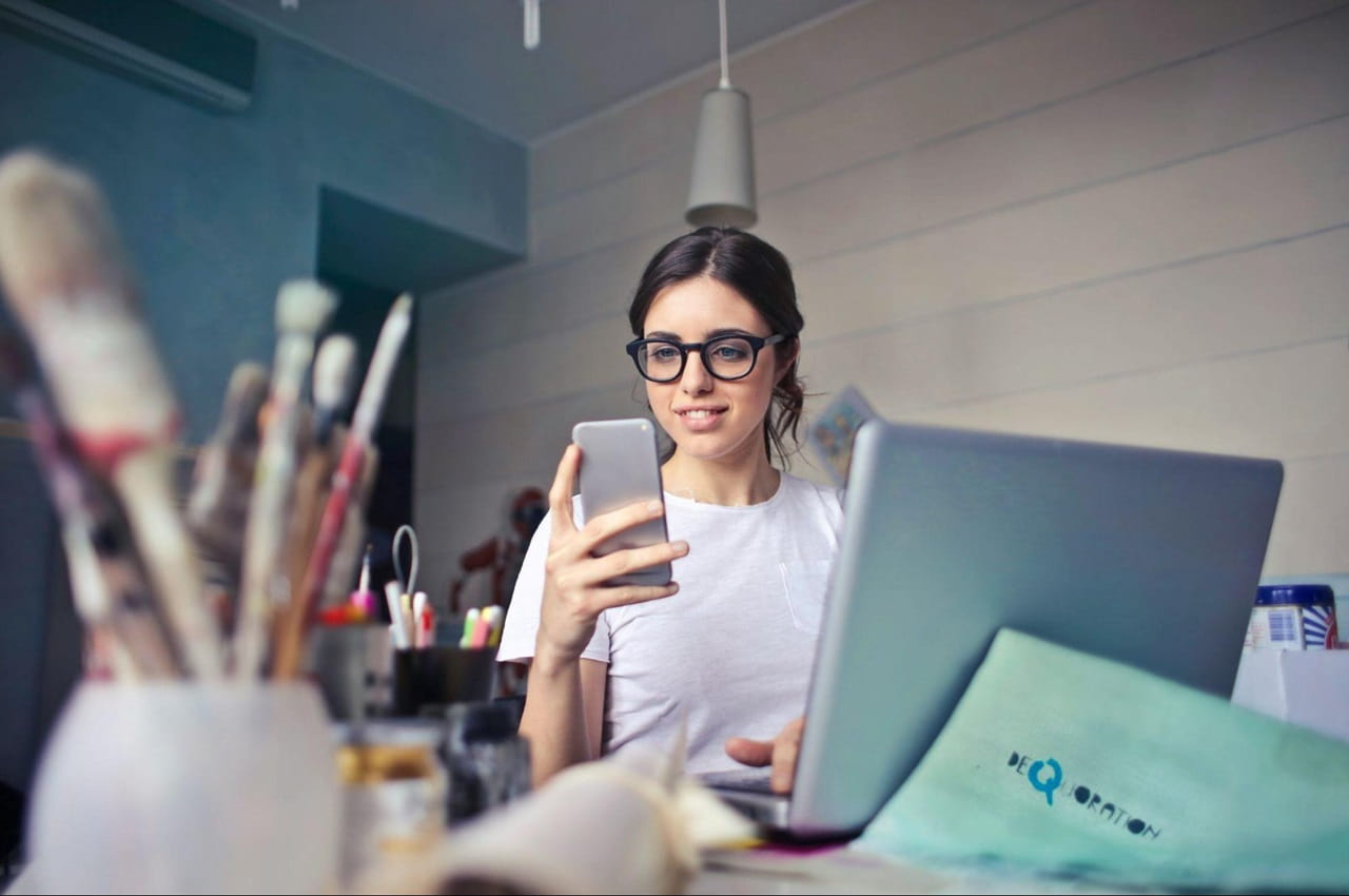 Mulher sorridente sentada em frente ao laptop segura um celular para acessar empréstimo pessoal BV. A mesa do escritório tem vários materiais, como lápis, caneta e caderno.