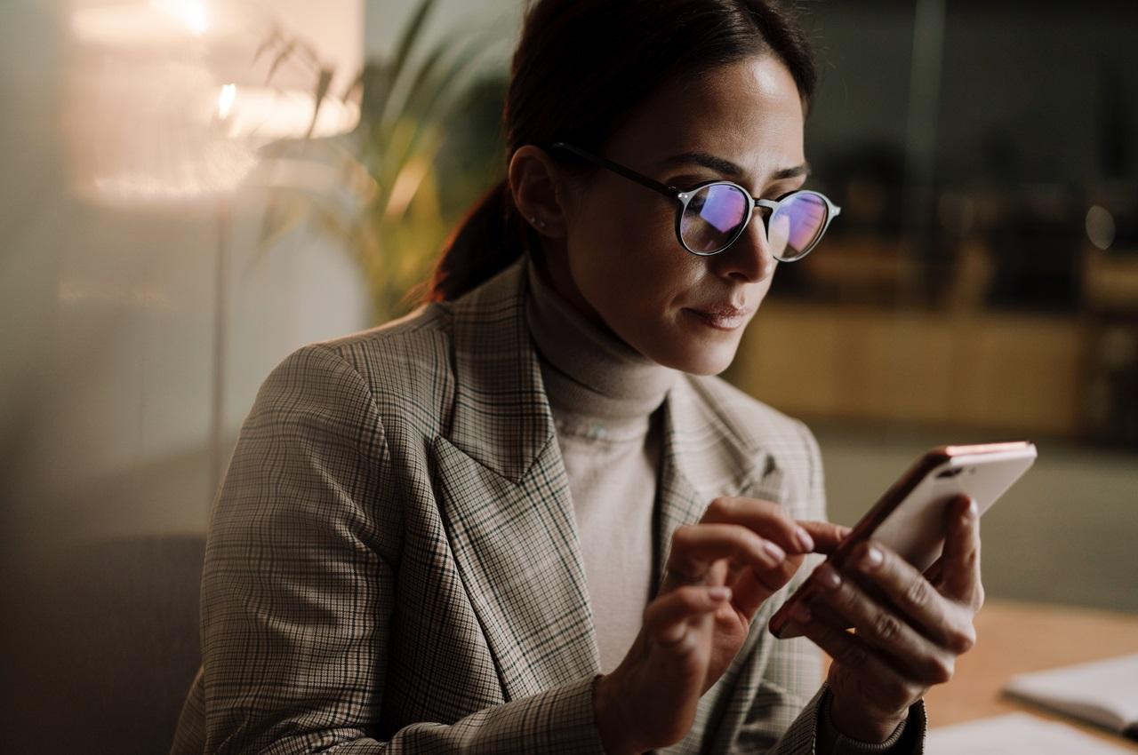 Mulher com expressão concentrada utilizando celular para consignado online. Ela está sentada no sofá de casa, usa óculos de grau e veste conjunto de alfaiataria.