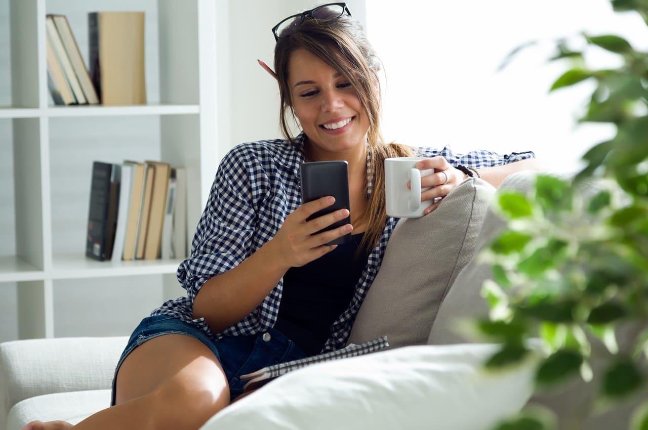 Jovem relaxando no sofá, sorrindo e segurando uma caneca enquanto olha para a tela do celular. Ela está pesquisando sobre empréstimo consignado público e privado.