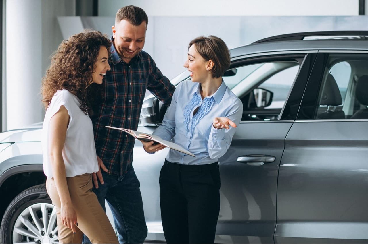 A imagem mostra um casal e a funcionária de uma concessionária, segurando uma caderneta, na frente de um carro.