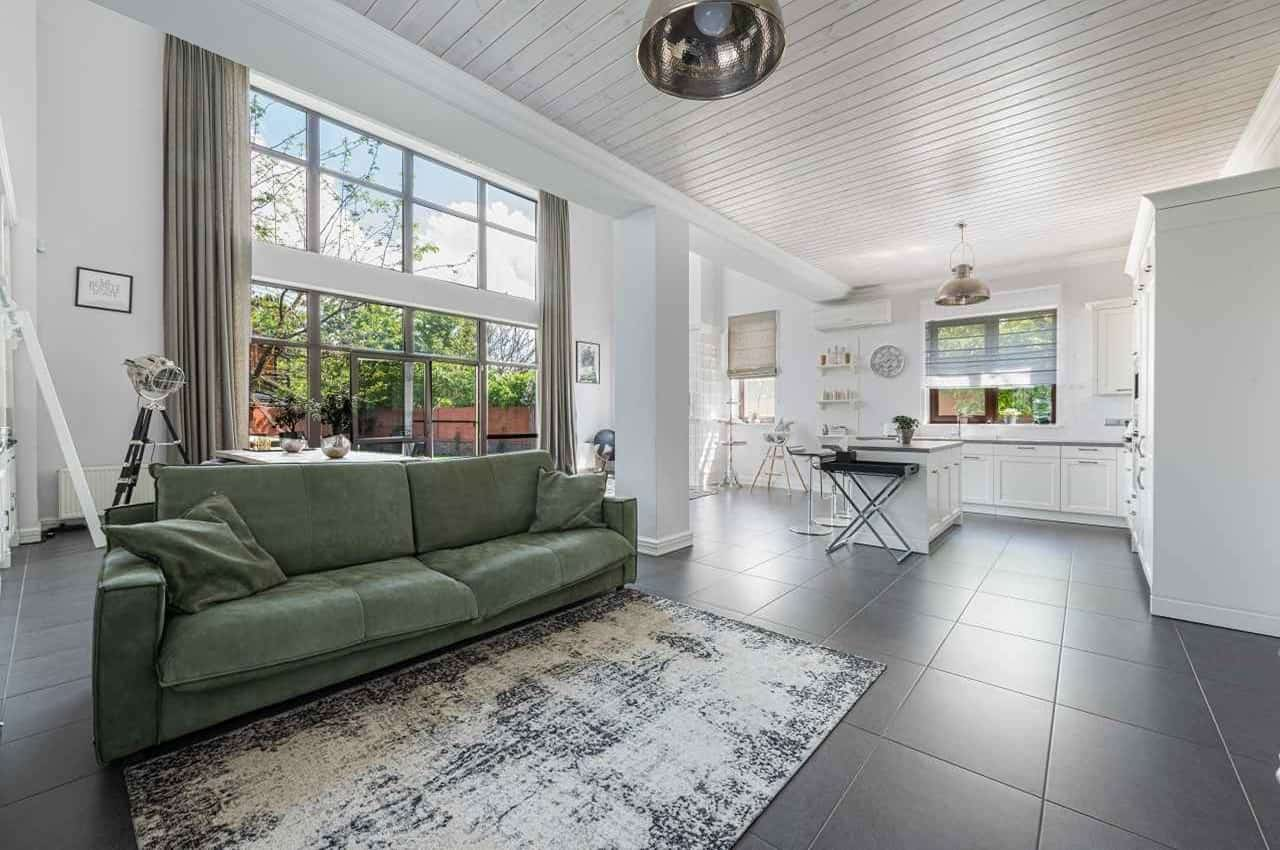 Sala de estar na cor branca e cozinha ao lado. A sala tem um sofá verde e tapete cinza e a cozinha eletrodomésticos e uma mesa de jantar branca.