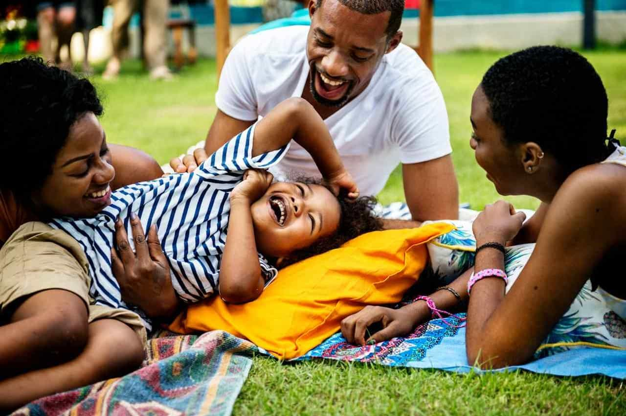 Família sorridente (homem, mulher e duas crianças pequenas) deitadas em uma toalha de piquenique na praça arborizada e o pai brinca com as crianças.