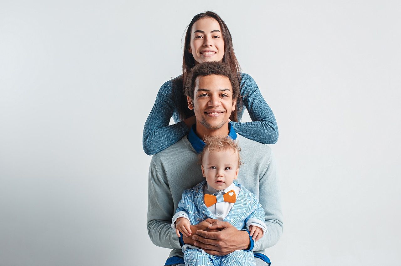 Família sorridente composta por uma mulher, um bebê pequeno e um homem. O homem está sentado no banco e segura o bebê no colo..