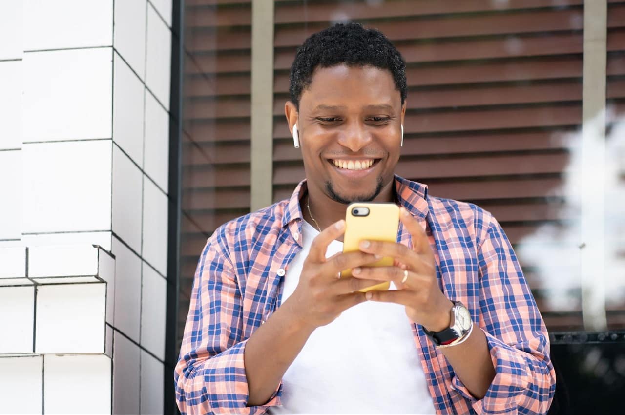 Homem sorridente, utilizando fones de ouvido e olhando para a tela do celular. Ele está parado em pé em uma rua movimentada.