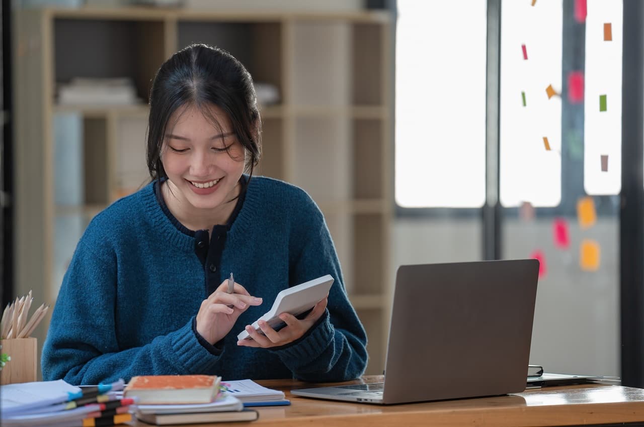 Mulher sorridente organizando documentações e contas na mesa, enquanto usa calculadora e um computador para declarar empréstimo no imposto de renda.