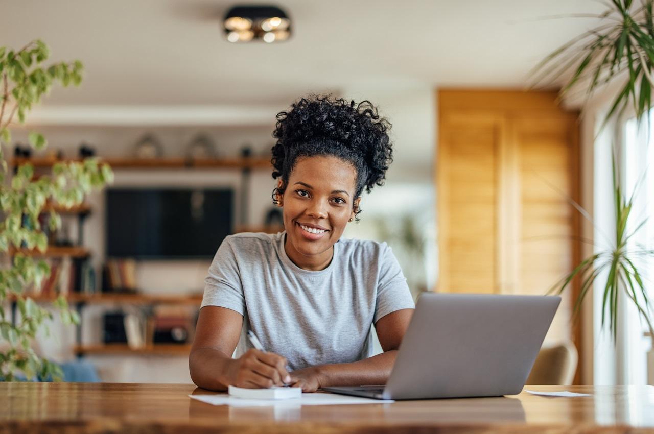 Mulher negra sorridente com cabelos presos e anotando em bloco de notas. Ela olha para frente e veste blusa básica cinza.