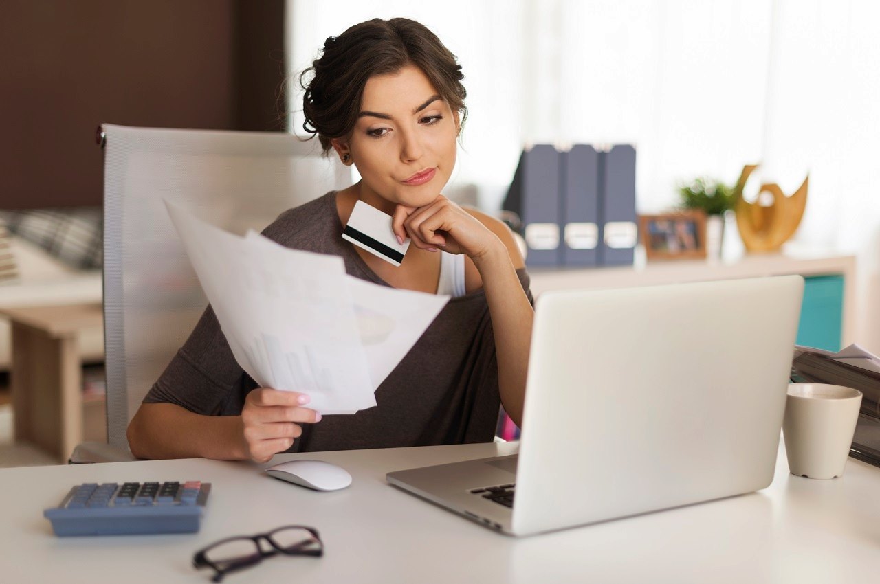 A imagem mostra uma mulher usando seu laptop e analisando alguns boletos. Ela está com seu cartão de crédito na mão e aparenta estar preocupada.