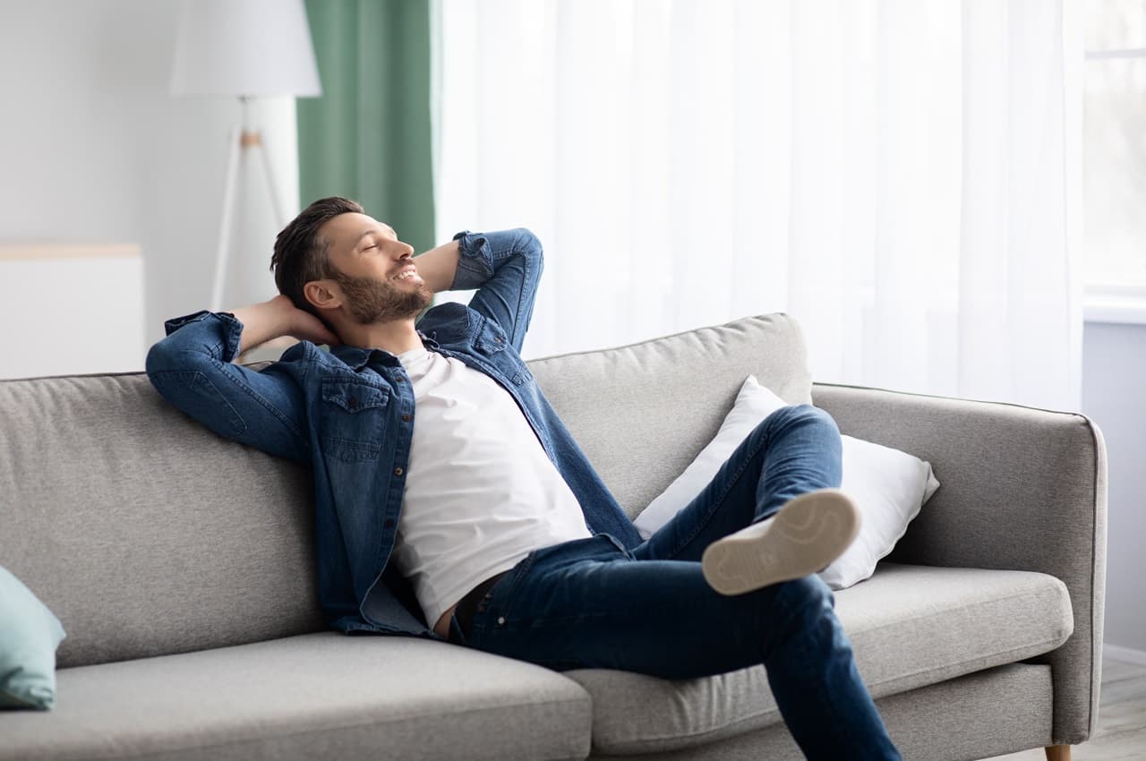 Homem sorridente com olhos fechados, sentado no sofá e com os braços esticados para trás. Ele veste calça jeans, blusa básica branca e uma jaqueta jeans.