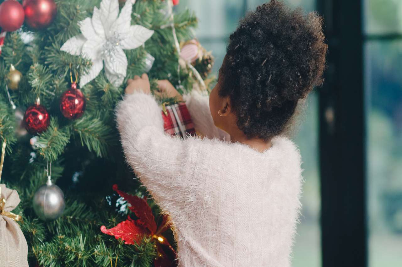 Criança pequena (ela está de costas na imagem) pendurando um enfeite em formato de caixa de presente na árvore de natal