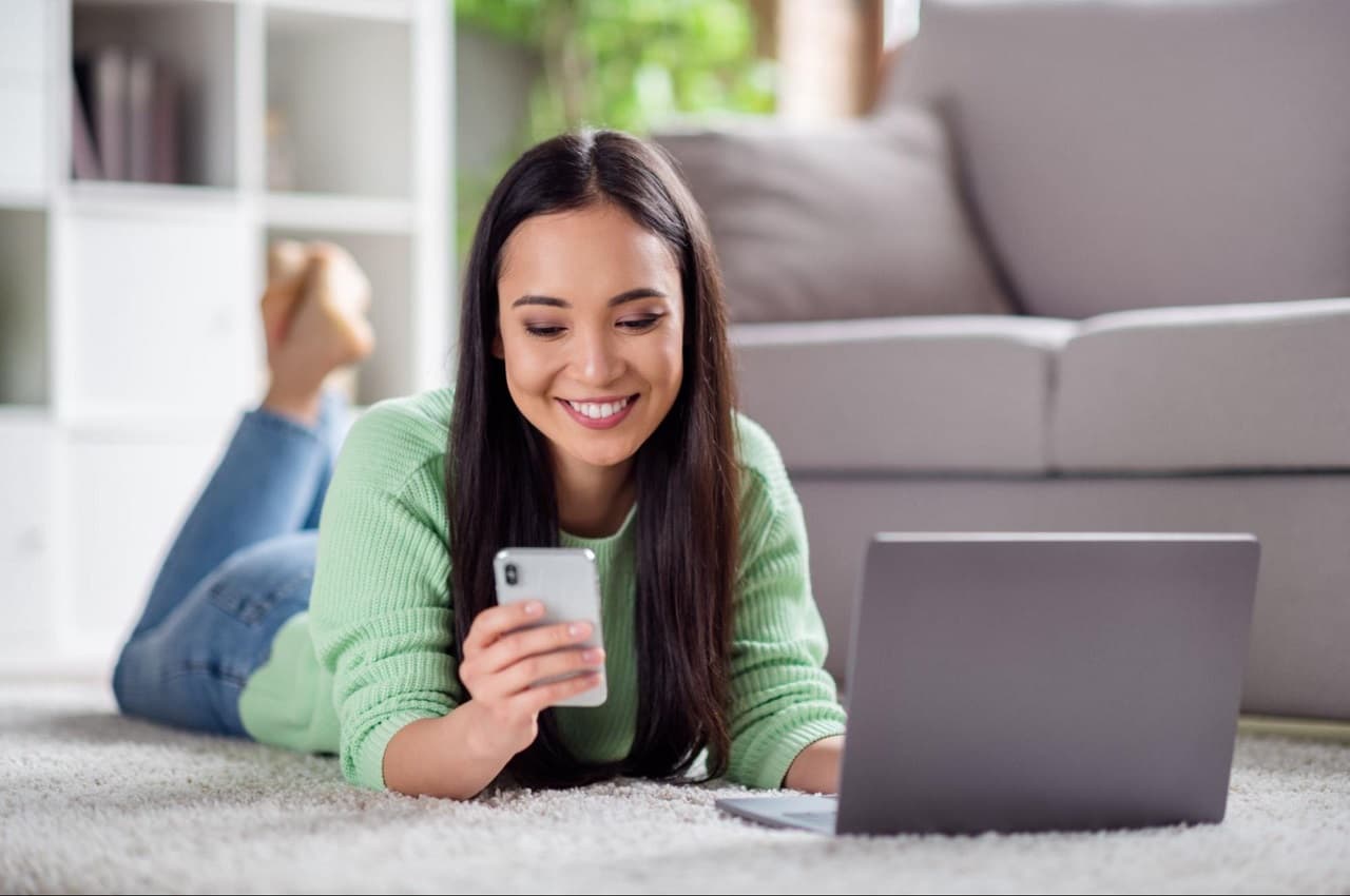 Mulher sorridente deitada de barriga para baixo no tapete da sala de estar e digitando no celular.