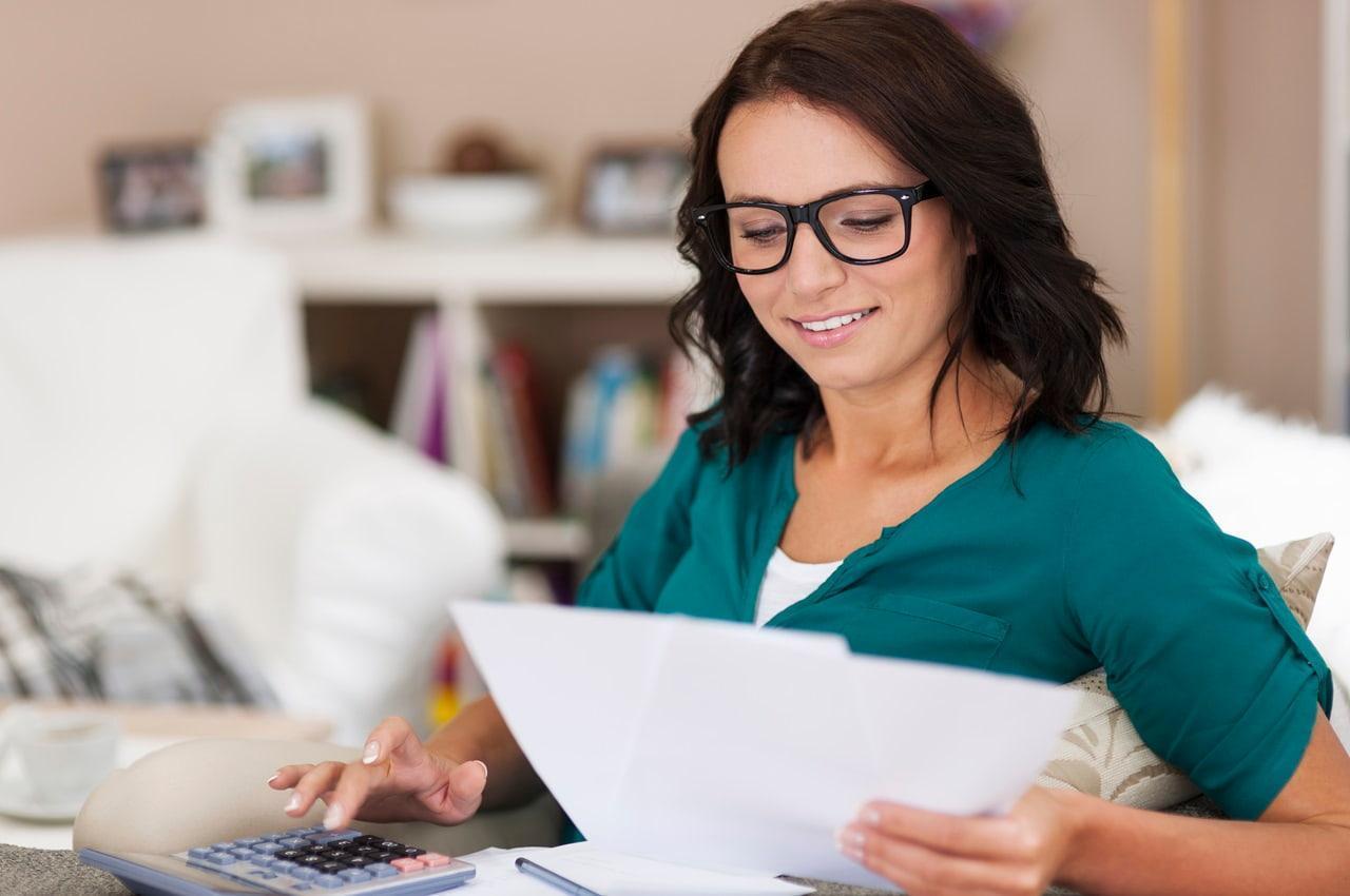 Mulher sorridente segurando alguns papéis em uma das mãos e utilizando uma calculadora com a outra. Ela está pagando as parcelas do seu empréstimo pessoal em dia.