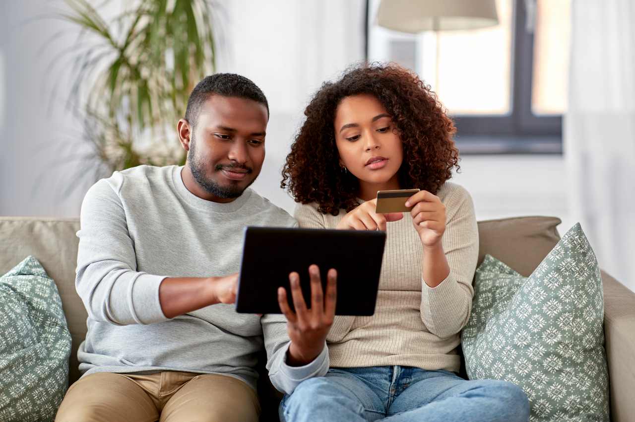 Homem segurando e apontando para um tablet, enquanto uma mulher sentada do seu lado confere um cartão de crédito.