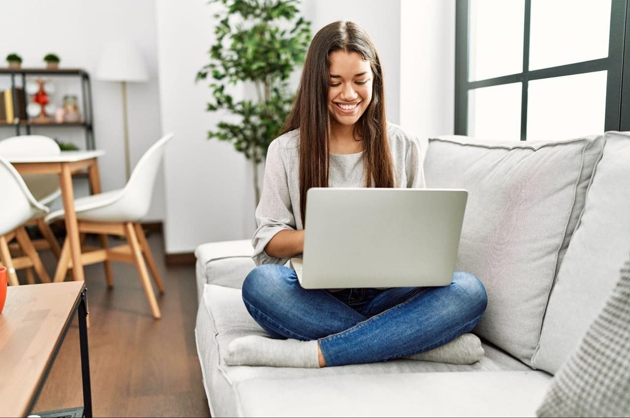 Mulher sorridente de cabelos castanhos longos, sentada de pernas cruzadas em cima do sofá e usando um laptop no colo. Ela veste blusa básica de mangas, calça jeans e meias. 