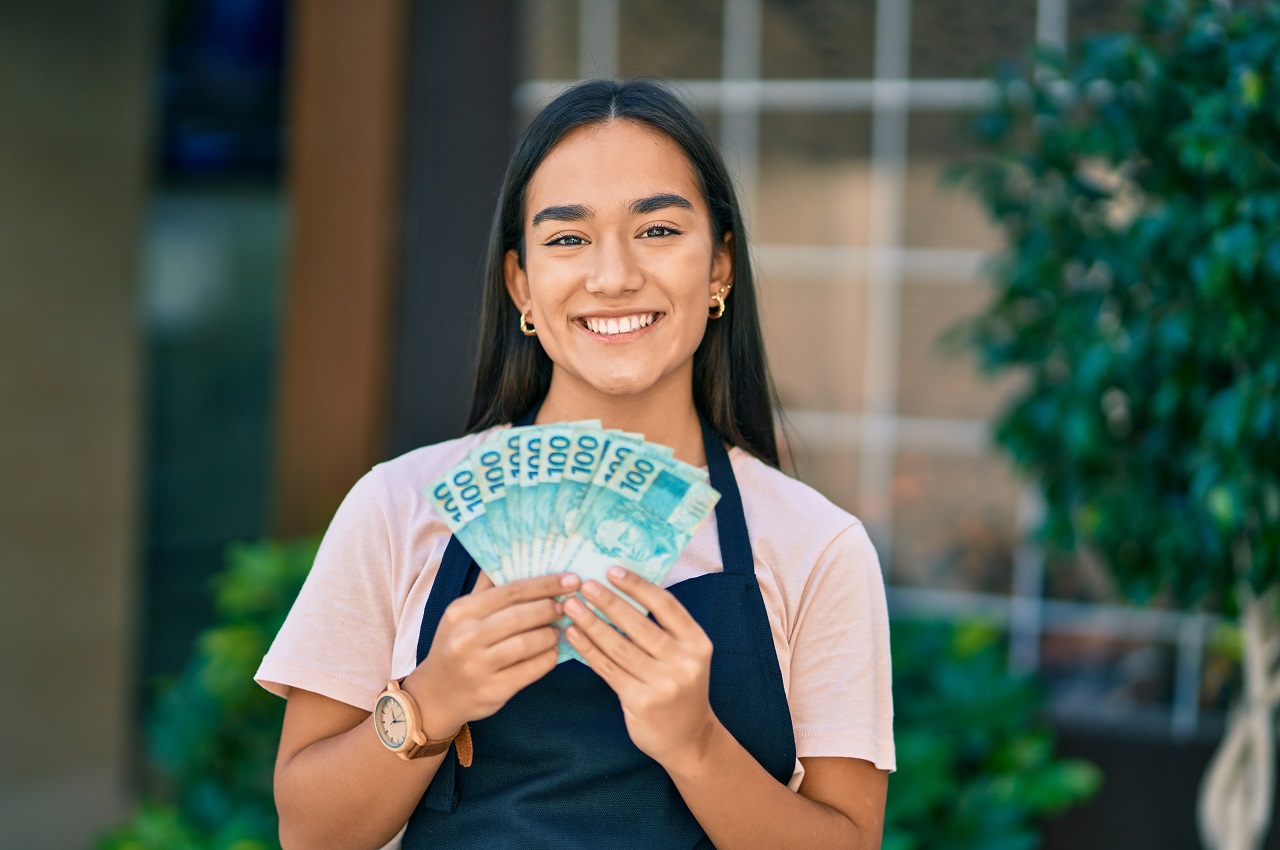 Mulher sorridente de cabelos longos, vestindo avental e segurando nas mãos um leque de notas de cem reais.