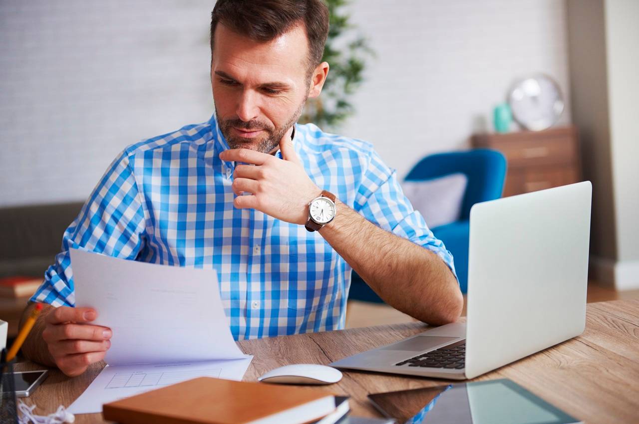 A imagem mostra um homem usando uma camisa xadrez, usando seu laptop e analisando um boleto. Ele aparenta estar preocupado.