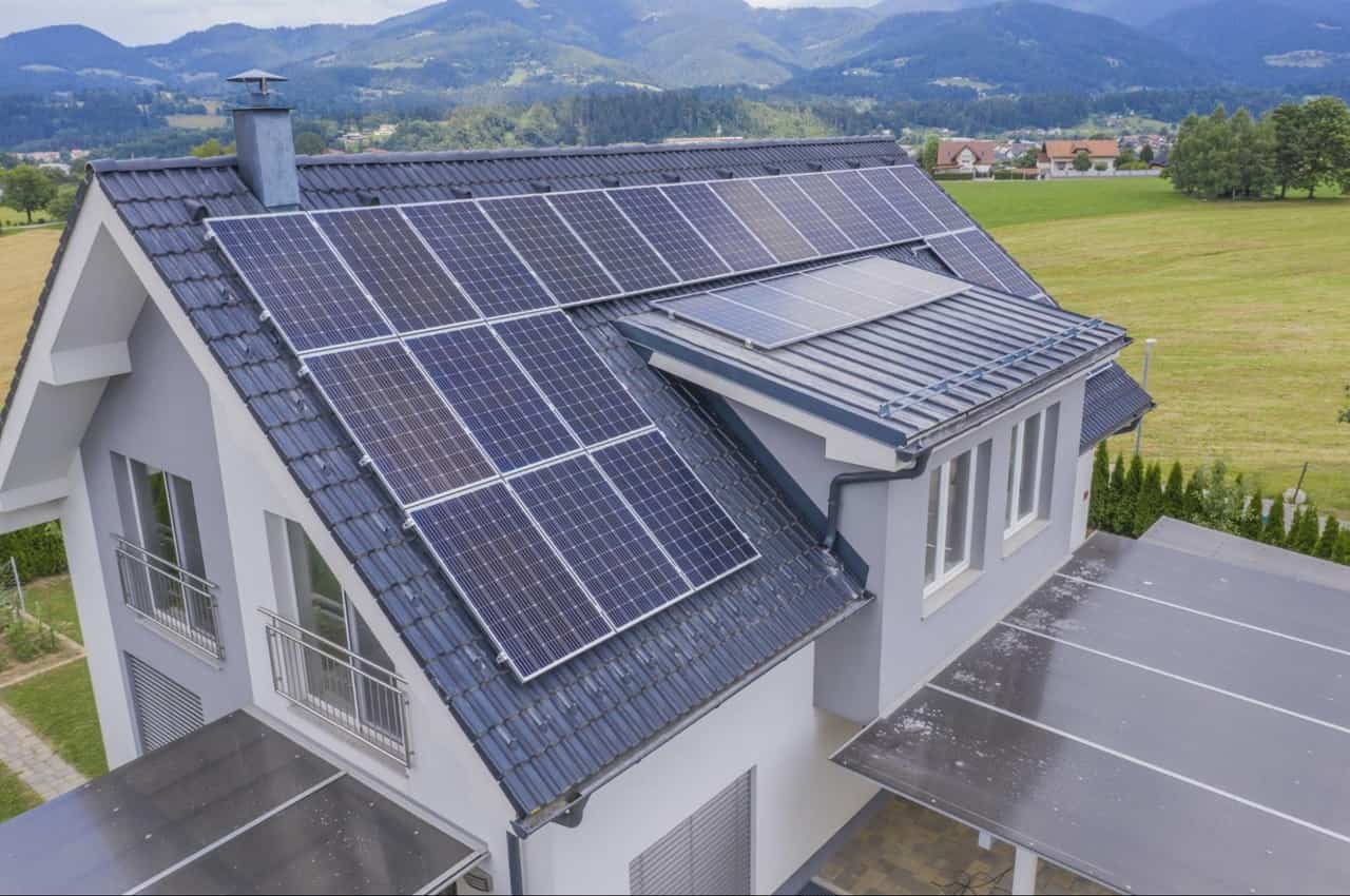 Casa de madeira com painéis solares no telhado, cercada por uma paisagem verde e montanhas ao fundo.