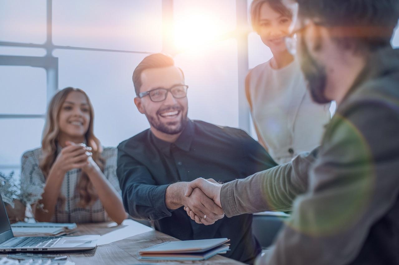 Grupo de pessoas sorridentes ao redor de uma mesa com computador, gráficos e cadernos. Na imagem, tem dois homens dando um aperto de mão enquanto duas mulheres observam.