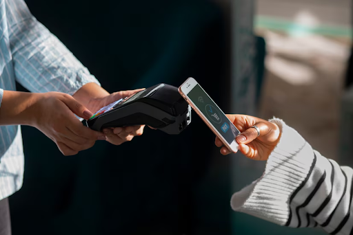 Uma mão segurando um terminal de pagamento enquanto outra mão aproxima um smartphone, indicando uma transação de pagamento por aproximação. O fundo é desfocado, destacando a ação de pagamento.