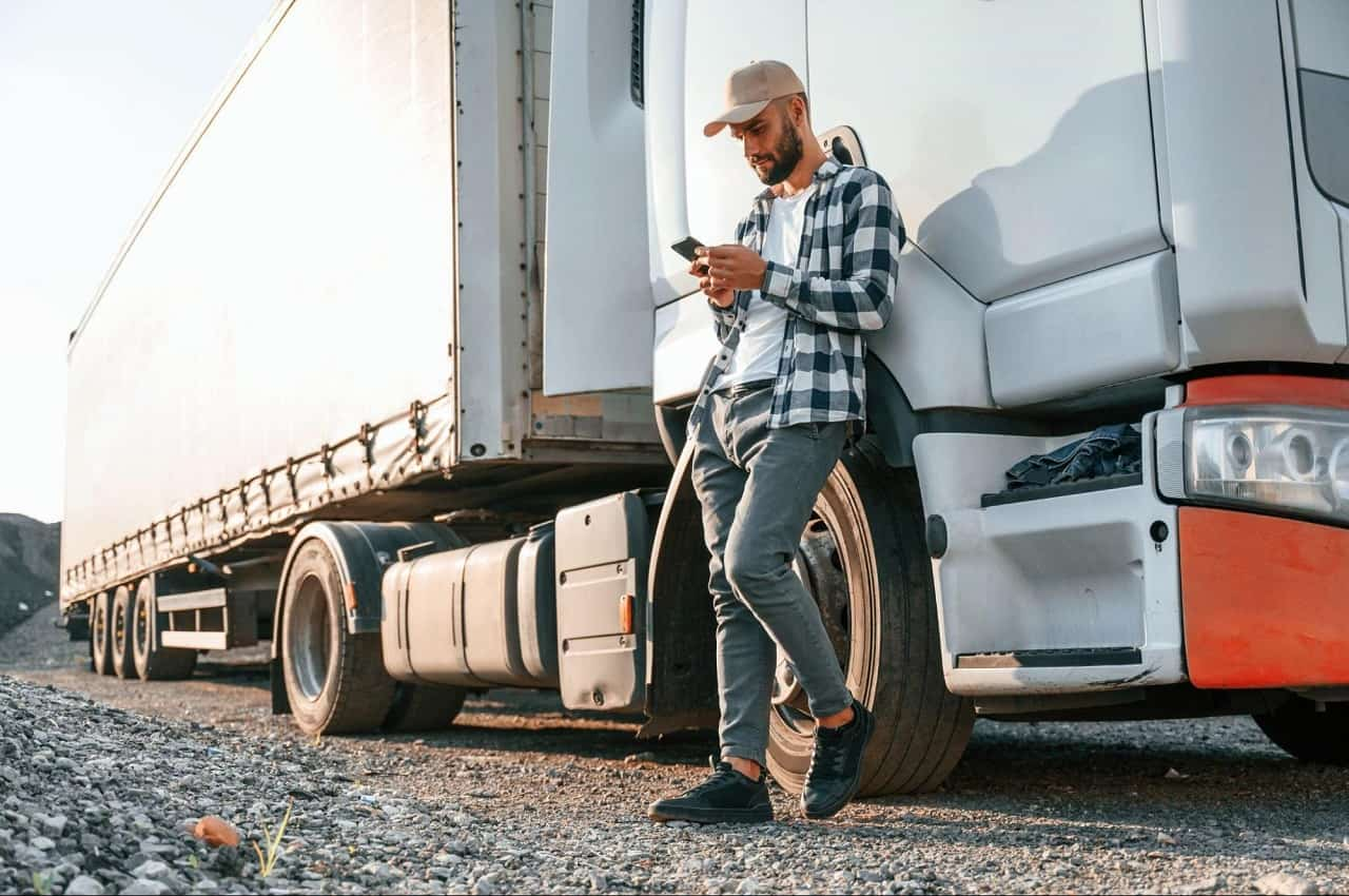 Homem de boné e blusa quadriculada, encostado em um caminhão vermelho e digitando no celular.