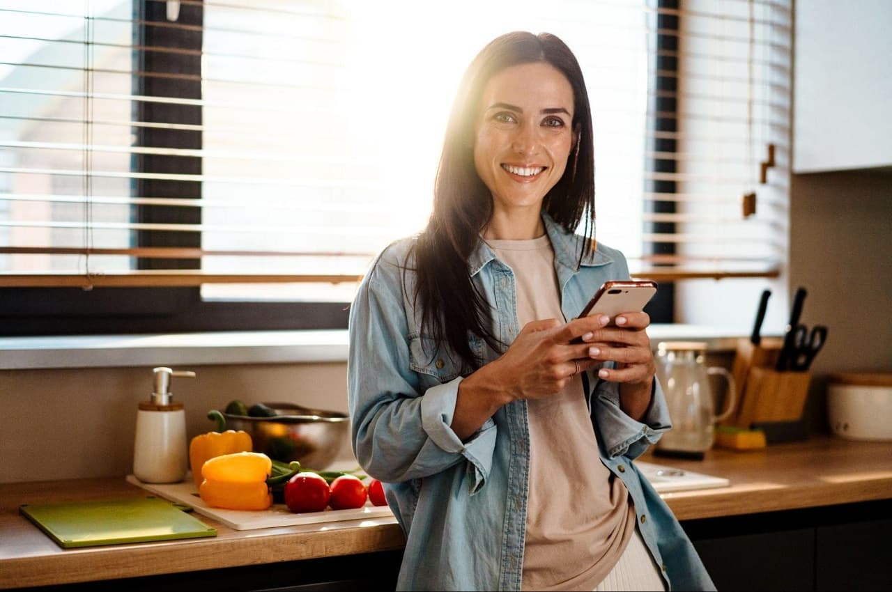 Mulher sorridente acessando dúvidas sobre empréstimo consignado no celular. Ela está encostada no balcão da cozinha e vestindo calça e jaqueta jeans